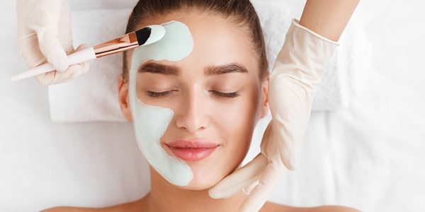 Beautician applying clay face mask on woman face, top view